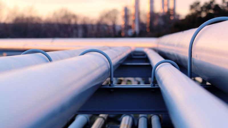 Nahaufnahme mehrerer parallel verlaufender metallischer Rohrleitungen einer Industrieanlage. Im unscharfen Hintergrund sind Industrieschornsteine im Licht der untergehenden Sonne zu sehen.