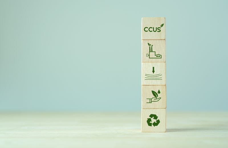 Übereinandergestapelte Holzwürfel mit der Aufschrift CCUS und Symbolen für Industrie, CO₂-Speicherung, Umweltschutz und Recycling.