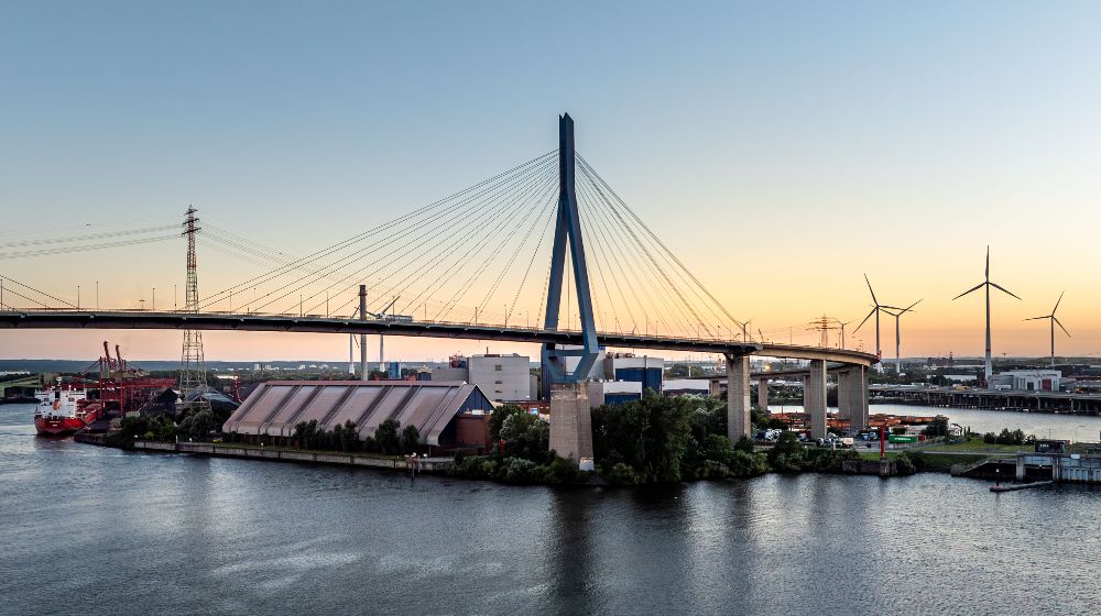Abendliche Ansicht einer Industrie- und Hafenlandschaft mit Schrägseilbrücke über einem Fluss; im Hintergrund stehen Windräder, Strommasten und Industrieanlagen.