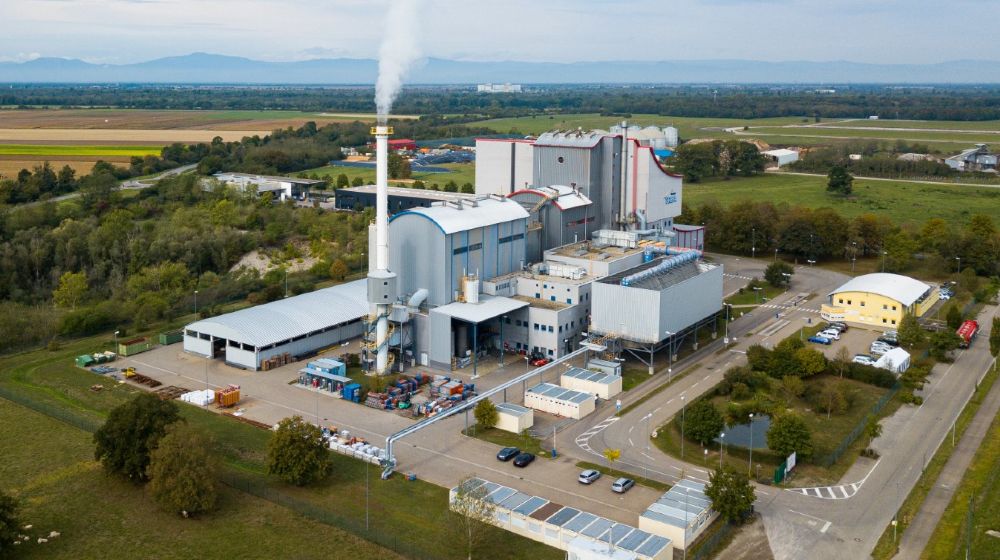 Luftaufnahme der Abfallverwertungsanlage TREA Breisgau mit Industriegebäuden und rauchendem Schornstein inmitten einer ländlichen Landschaft.